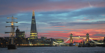 London Tower Bridge Shard sailing ship This urban landscape photograph captures the city of London in the United Kingdom during an evening sunset in early autumn. The main subjects of the image include the iconic Tower Bridge, the modern Shard skyscraper, and a sailing ship, all prominent within the city’s architecture. The illuminated bridge stretches across the River Thames, with the Shard glowing in the urban skyline, while dramatic sunset colors highlight the buildings and sky. The photograph’s composition showcases London’s blend of historic and contemporary architecture as the evening light casts a warm glow over the city.